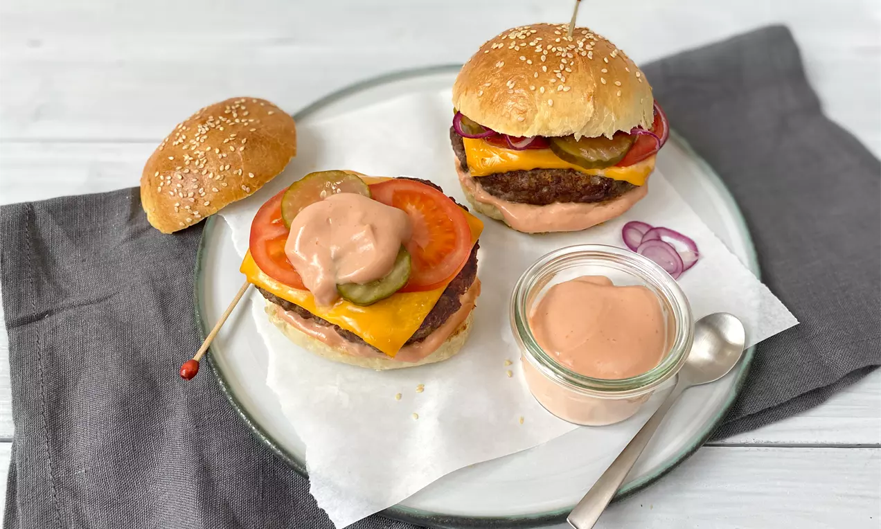 Burgerbrötchen mit Sesam, rote Zwiebeln und saftig belegte Burger mit geschmolzenem Käse neben einem Glas mit cremiger rosa Soße