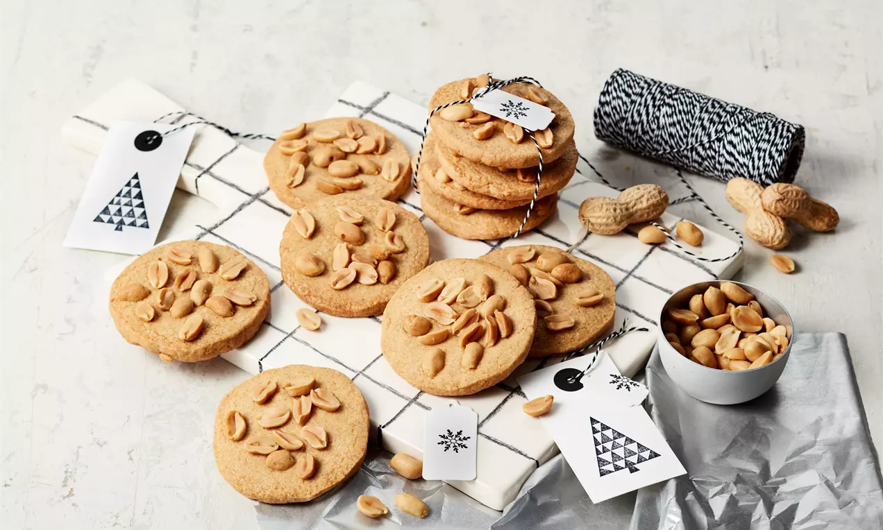 Goldbraune Erdnussplätzchen mit gerösteten Erdnüssen, flach und rund, auf einem karierten Tuch, daneben Nüsse und kleine Geschenkanhänger