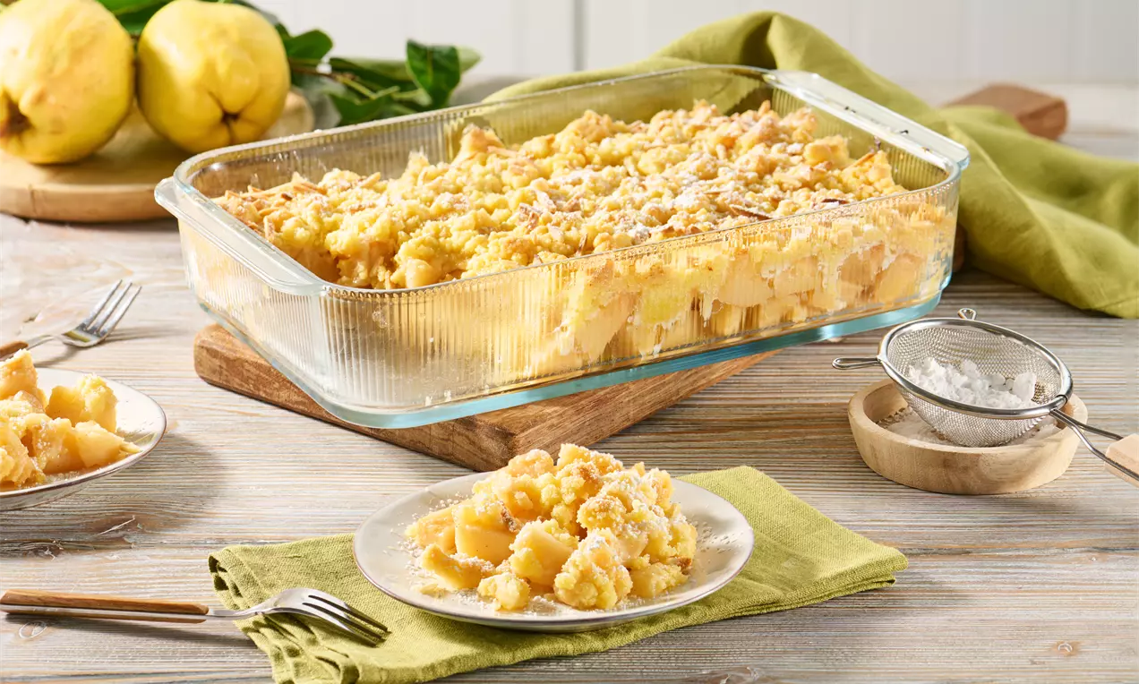 Goldbrauner Quitten-Crumble mit knusprigen Streuseln in einer eckigen Glasform, serviert auf Tellern mit fruchtigen Stücken und Puderzucker