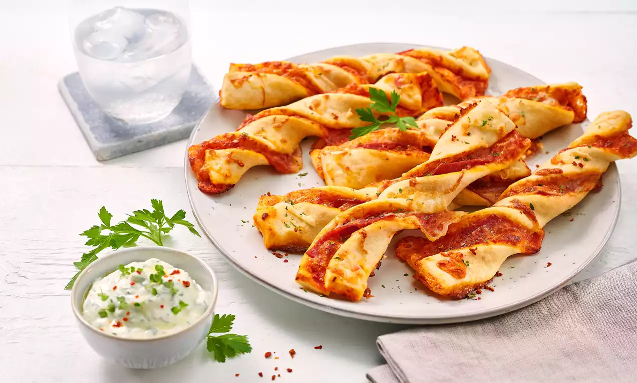 Gedrehte, goldgelbe Pizzastangen mit roten Tomatensaucestreifen und grünen Kräutern, serviert auf weißem Teller mit Dip und Wasser im Glas