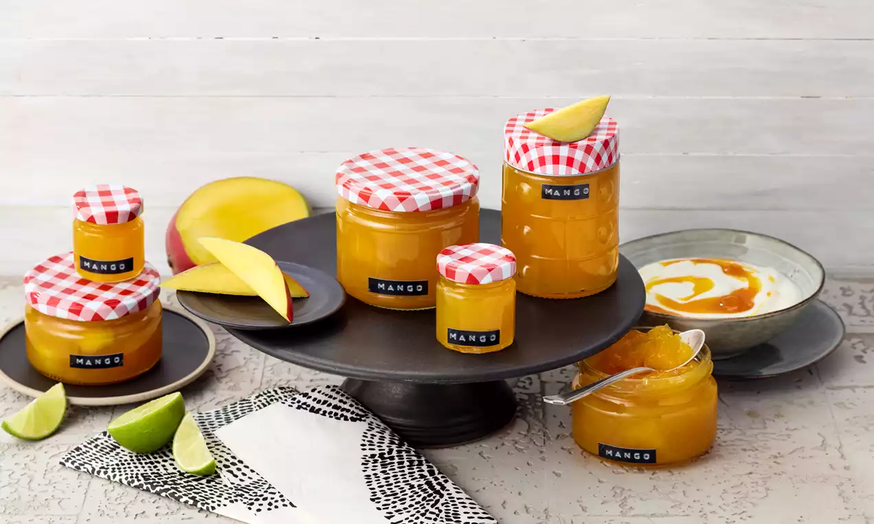 Gläser mit goldgelber Mango-Marmelade und rot-weiß karierten Deckeln auf schwarzer Etagere, daneben Mango, Limetten und Marmelade auf Teller