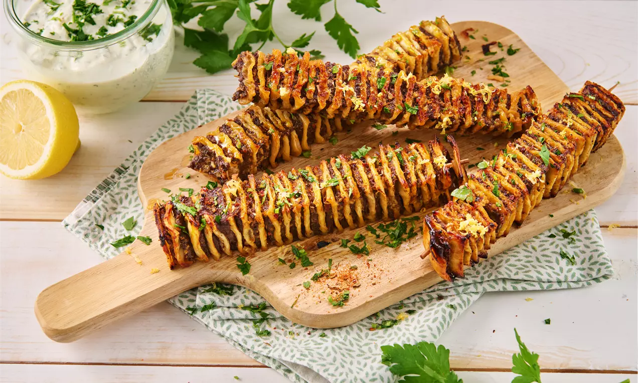 Goldbraune Kebabspieße mit spiralartiger Textur, garniert mit frischen Kräutern, auf einem hölzernen Brett mit Zitronenscheibe und Joghurt-Dip