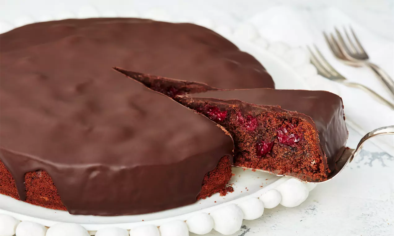 Schoko-Kirsch-Kuchen mit glänzendem dunklen Guss, angeschnittener brauner Rührteig mit sichtbaren roten Kirschen auf weißer Platte