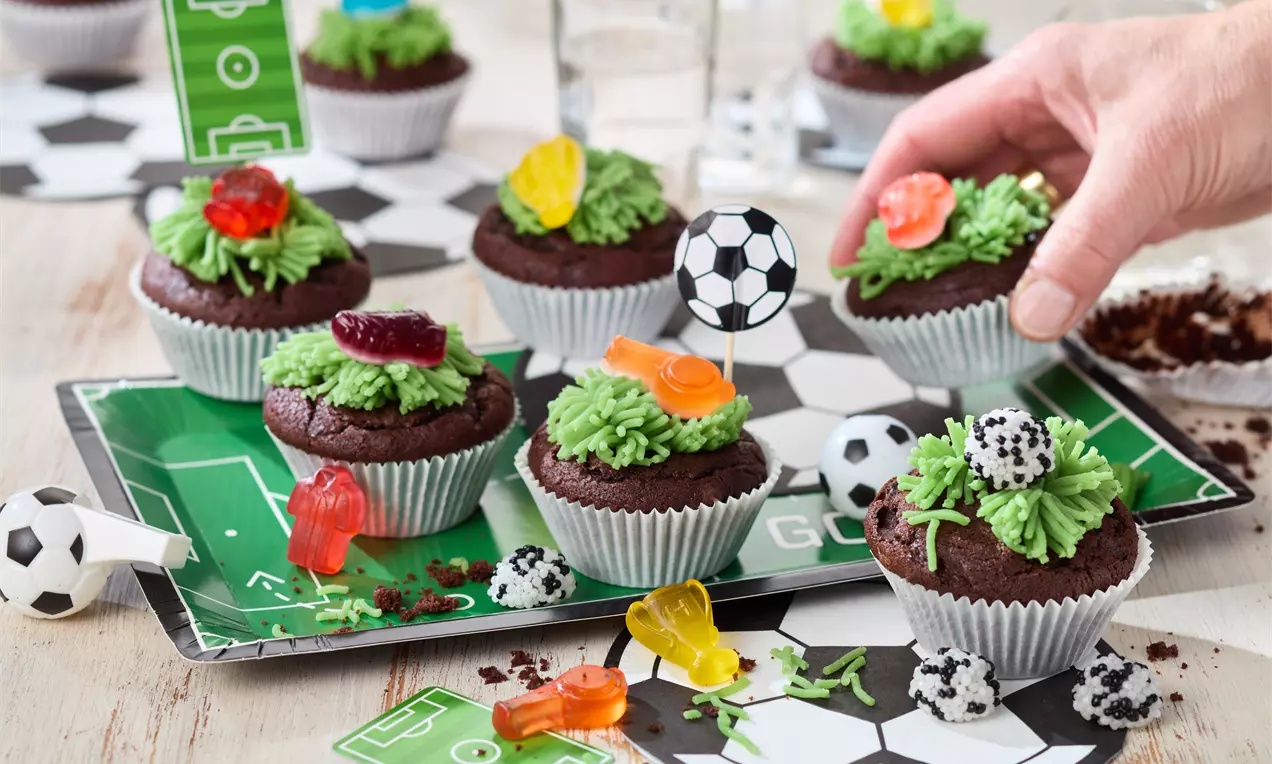 Schokoladen-Muffins mit grüner Creme verziert, Fußball-Dekorationen und bunten Gummibärchen auf Fußball-Muster serviert, Hand greift nach einem