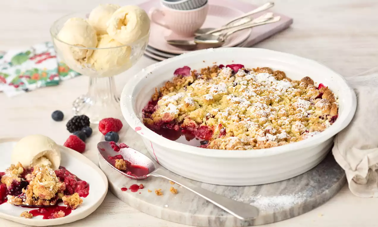 Goldbrauner Beeren-Crumble mit saftigen roten und schwarzen Beeren, bestäubt mit Puderzucker in einer weißen Form auf Marmorplatte