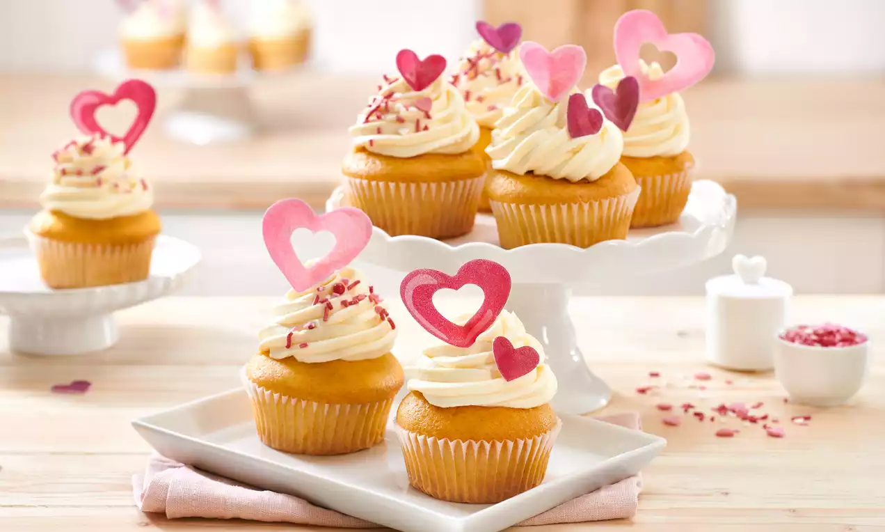 Hochzeitsmuffins mit goldgelbem Teig, weißer Sahnehaube und rosafarbenen Herzdekoren auf weißem Geschirr liebevoll angerichtet
