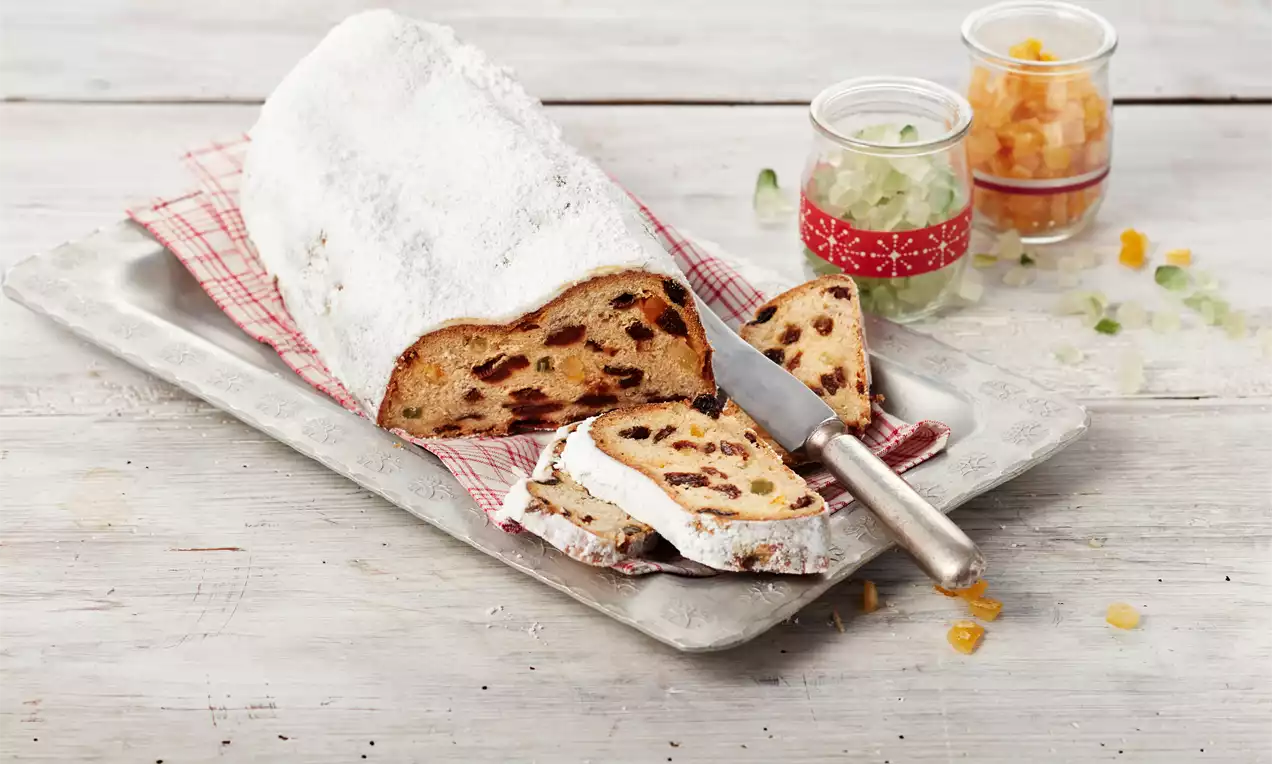 Ein goldbrauner Butterstollen mit Puderzuckerschicht, angeschnitten auf einem Tablett mit rotem Tuch, dekoriert mit kandierten Früchten