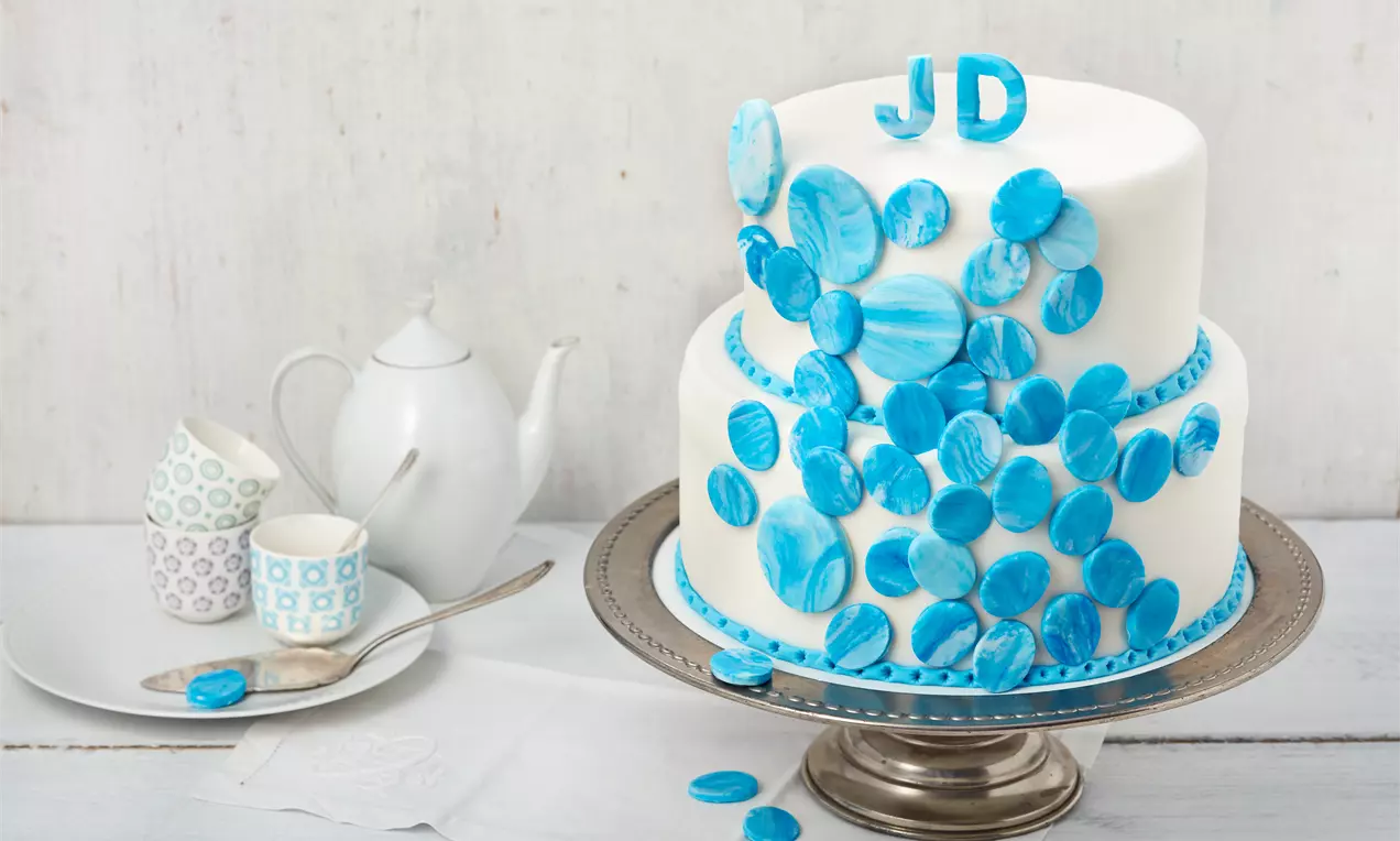 Zweistöckige weiße Hochzeitstorte mit blauen marmorierten Dekorplättchen und Initialen JD auf silberner Platte neben Porzellantassen und Teekanne