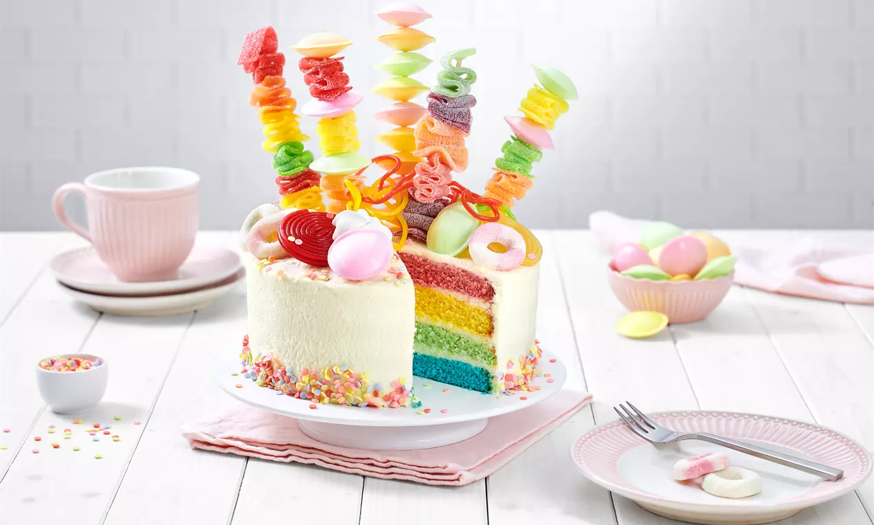 Bunte Regenbogentorte mit weißer Creme und Zuckerstreuseln, verziert mit bunten Süßigkeitenspießen auf rosa Tischdecke angerichtet