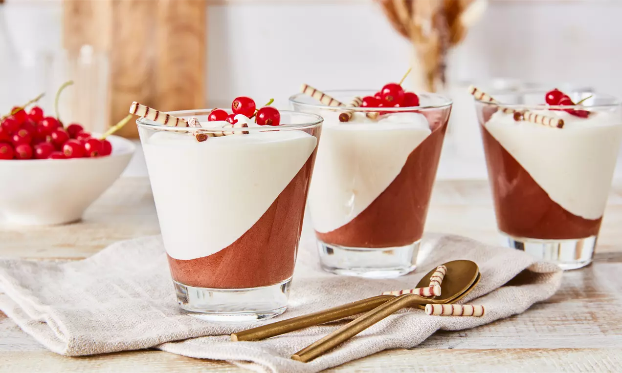 Schwarz-Weiße-Mousse in klaren Gläsern mit diagonal getrennter weißer und brauner Creme, dekoriert mit roten Johannisbeeren und Schokostäbchen