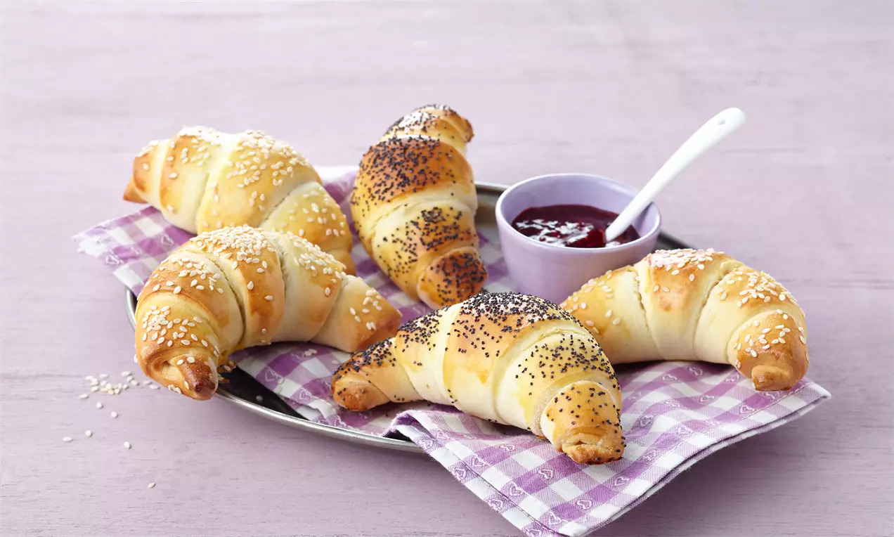 Goldgelb gebackene, filigrane Hörnchen mit Mohn und Sesam bestreut auf kariertem Tuch, dazu eine violette Schale mit roter Marmelade