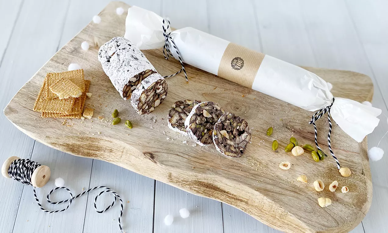 Schoko-Salami mit weißem Puderzucker, angeschnitten auf einem rustikalen Holzbrett, daneben Haselnüsse, Pistazien und Butterkekse