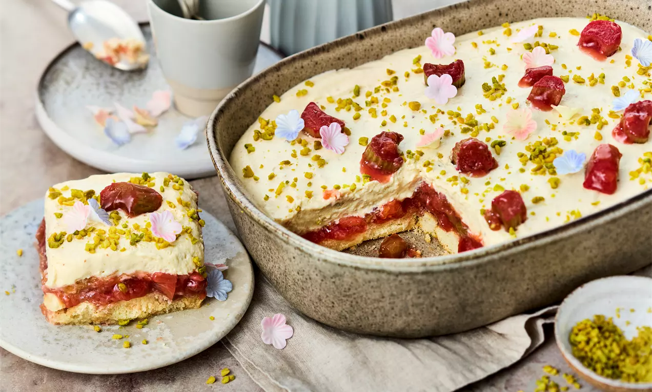 Rhabarber-Tiramisu in einer rustikalen Form, mit Schichten aus hellem Mascarpone, rotem Kompott und Löffelbiskuits, garniert mit Pistazien und Blüten