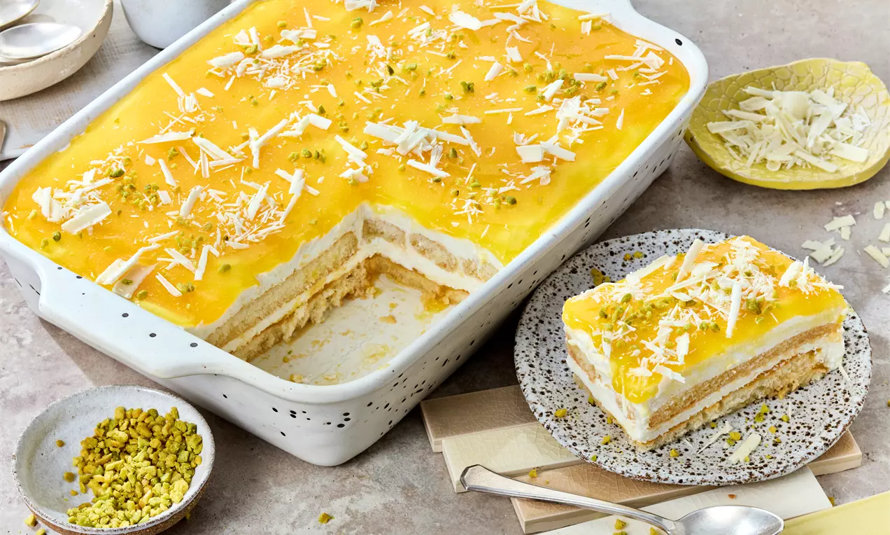 Solero-Tiramisu mit gelber Fruchtschicht, weißen Schokoraspeln und Pistaziensplittern in einer weißen Auflaufform, daneben ein Stück auf einem Teller