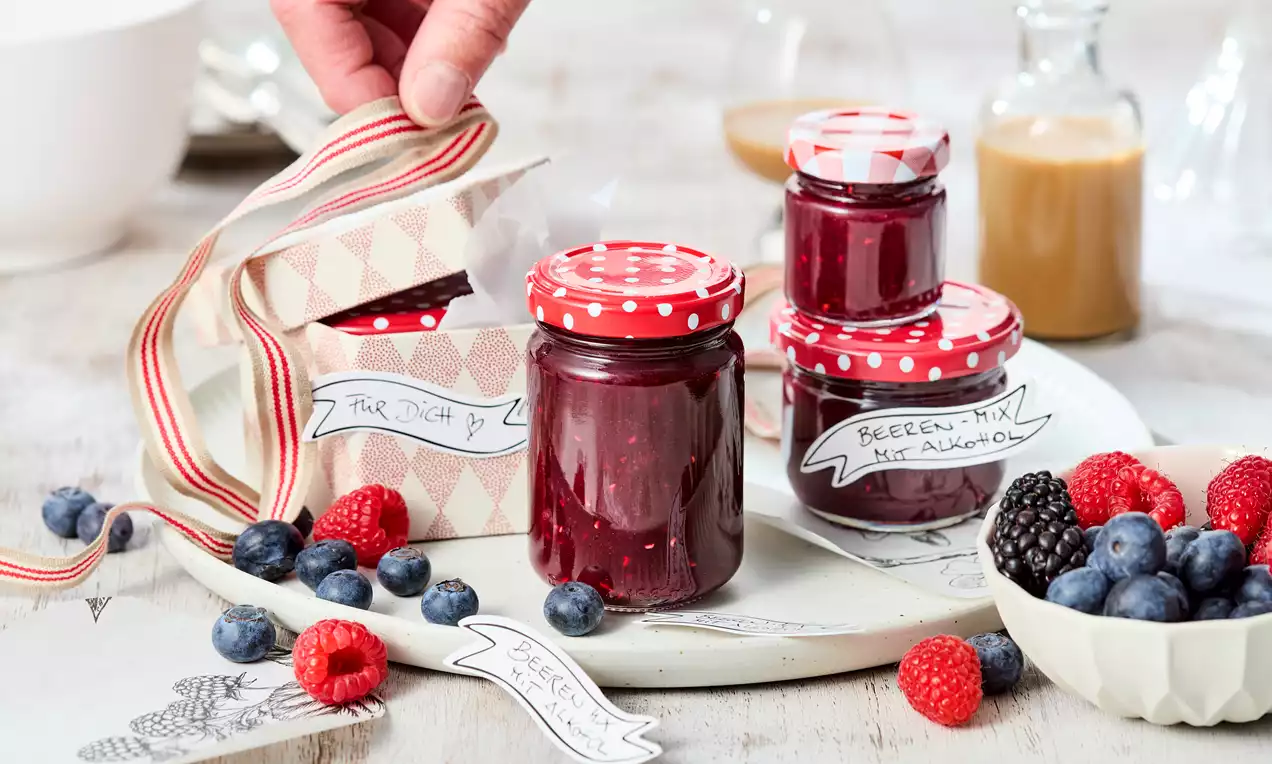 Drei Glasbehälter mit roter Marmelade und gepunkteten Deckeln auf hellem Holz, dekoriert mit Beeren und Etiketten sowie einer Stoffschleife