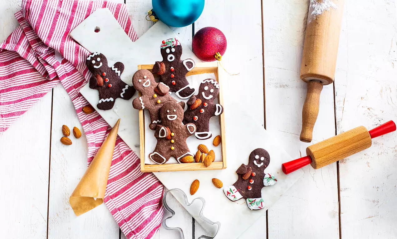 Lebkuchenmännchen aus dunklem Teig mit Zuckerdekoration auf einem Holztablett, umgeben von Mandeln, Teigrolle und weihnachtlicher Deko