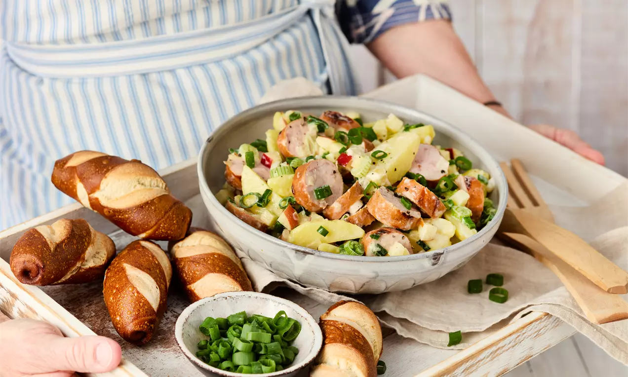 Bratwurstsalat mit zartbraunen Wurstscheiben, Kartoffeln und frischen Kräutern in einer grauen Schale auf einem Holztablett mit Laugenstangen