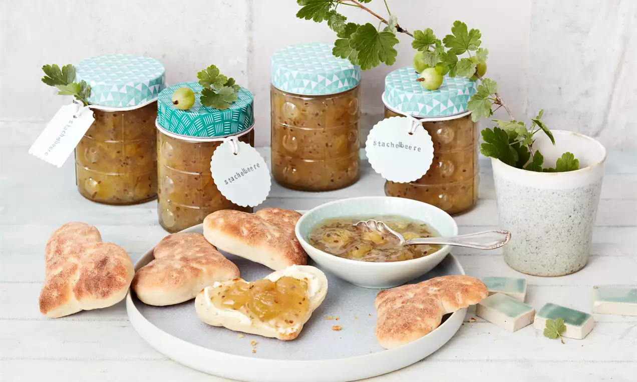 Stachelbeermarmelade in offenem Glas, leuchtend gelbgrün mit sichtbaren Fruchtstücken, Vanilleschote und frischen Beeren, Löffel auf Brett