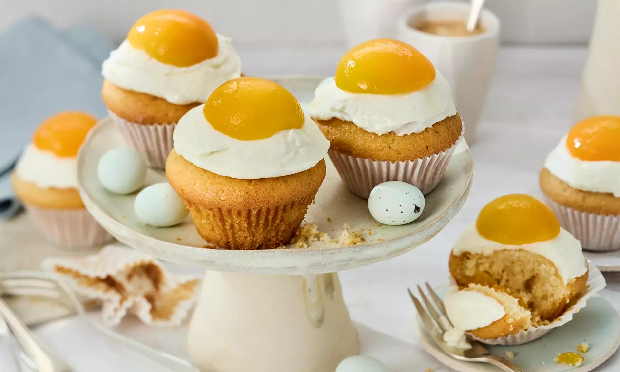 Goldbraune Muffins mit weißer Creme und glänzenden gelben Aprikosenhälften auf einer Keramiketagere, umgeben von kleinen hellblauen Eiern