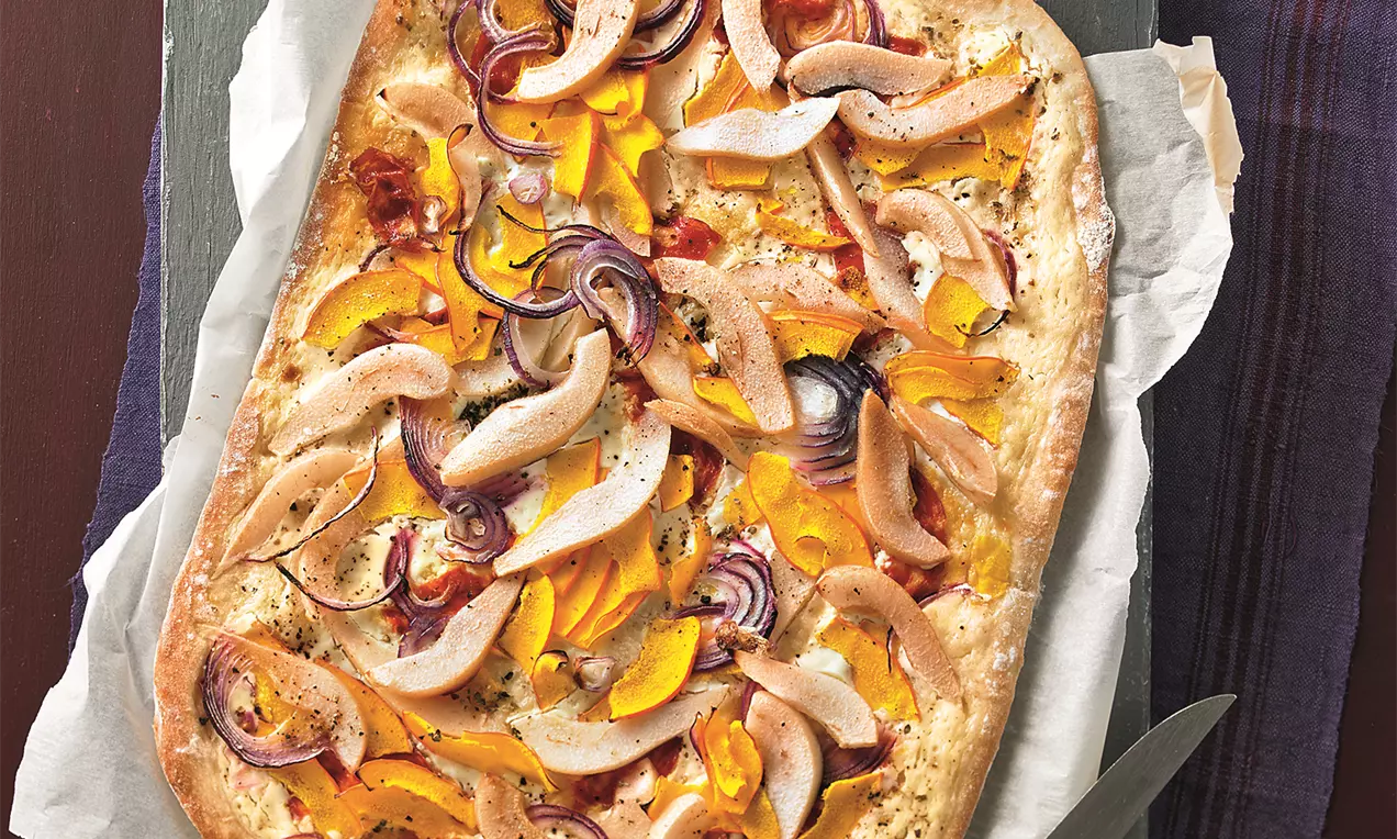 Rechteckiger Flammkuchen mit dünnen Kürbis- und Birnenscheiben, roten Zwiebeln und Kräutern auf heller Backunterlage angerichtet
