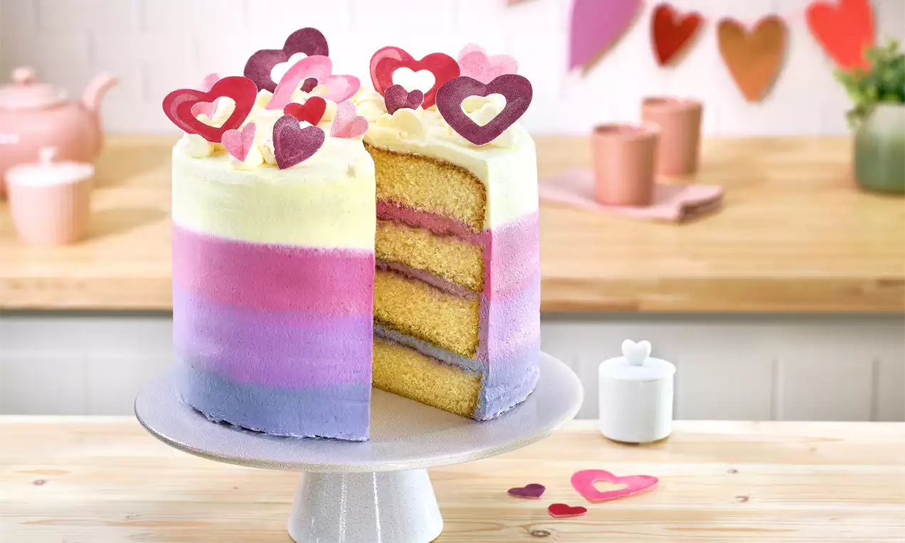 Hohe, pastellfarbene Torte mit lila, rosa und weißer Creme, geöffnet mit gelben Schichten, dekoriert mit Herzornamenten auf einer Tortenplatte