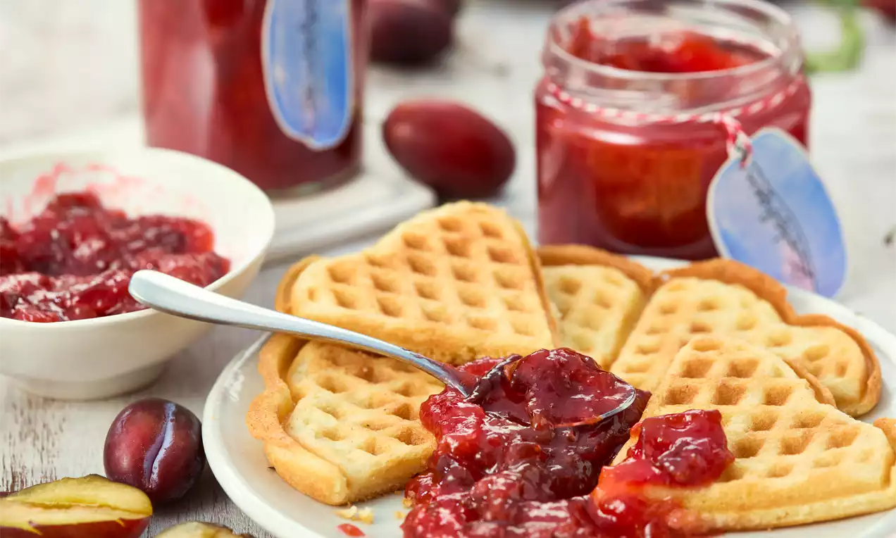 Goldbraune, herzförmige Waffeln auf einem Teller, bestrichen mit glänzend roter Pflaumengrütze, daneben offene Gläser und frische Pflaumen
