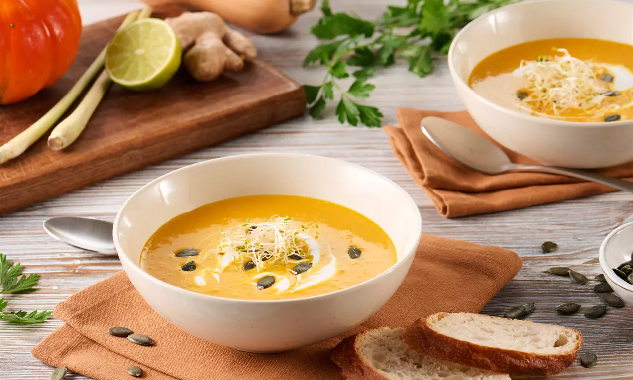 Cremige orangefarbene Kürbissuppe mit glänzender Oberfläche, garniert mit hellen Sprossen und Kürbiskernen in einer weißen Schale auf einem Stoffset