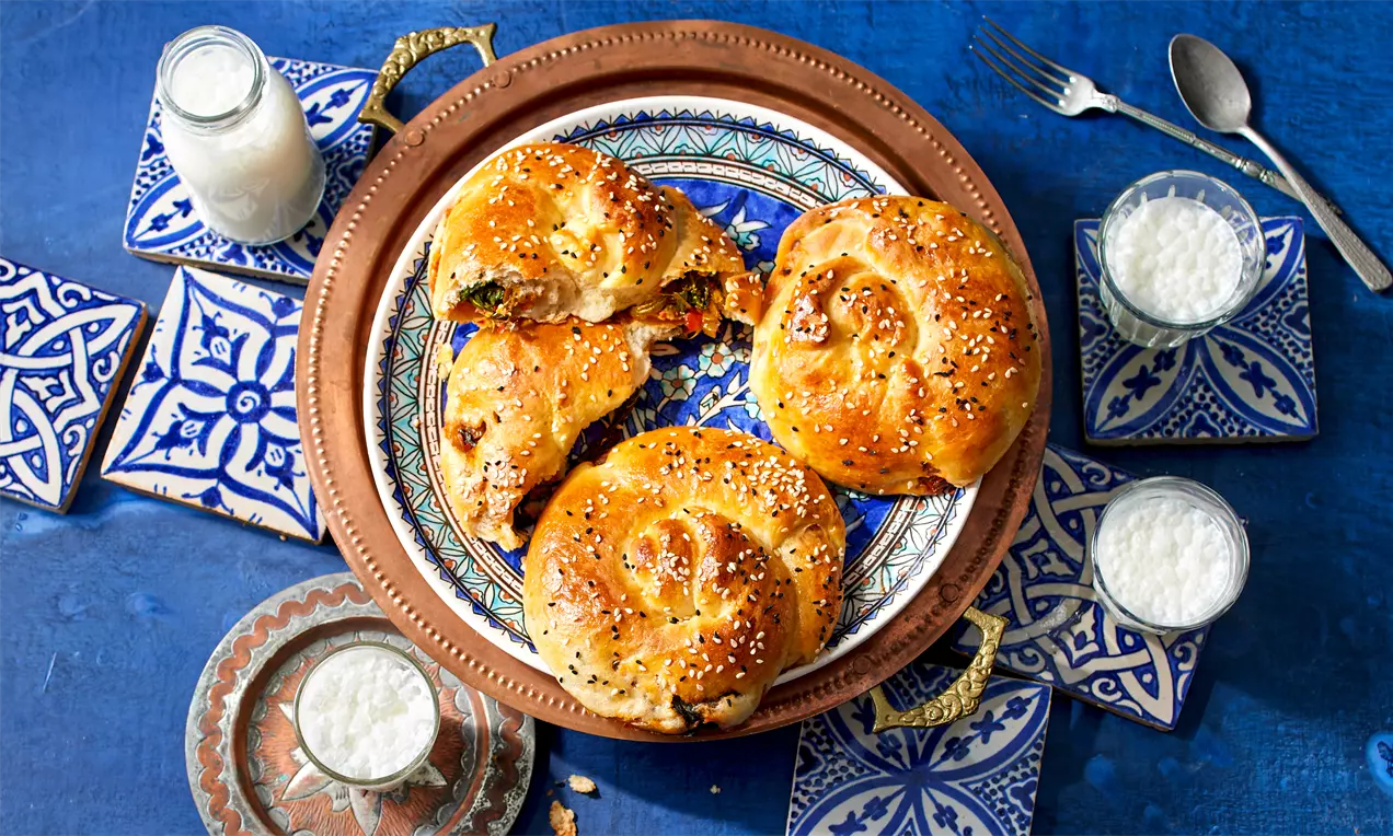 Goldbraune Börek mit Spinatfüllung, sesam-bestreut auf gemustertem Teller, arrangiert mit kleinen Gläsern Joghurt auf blauem Untergrund