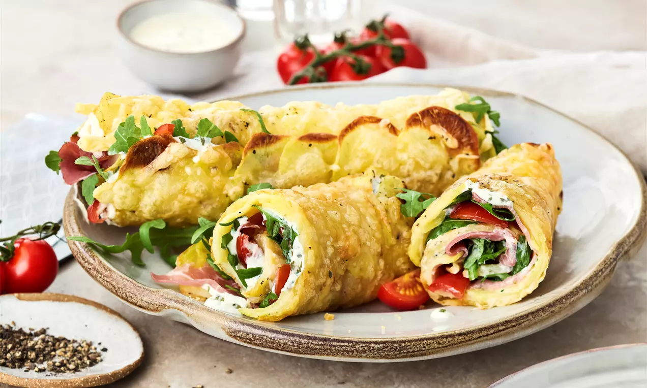 Goldene Kartoffel-Wraps mit knuspriger Textur, gefüllt mit roten Tomaten, grünem Rucola und belegtem Schinken auf einem hellen Teller