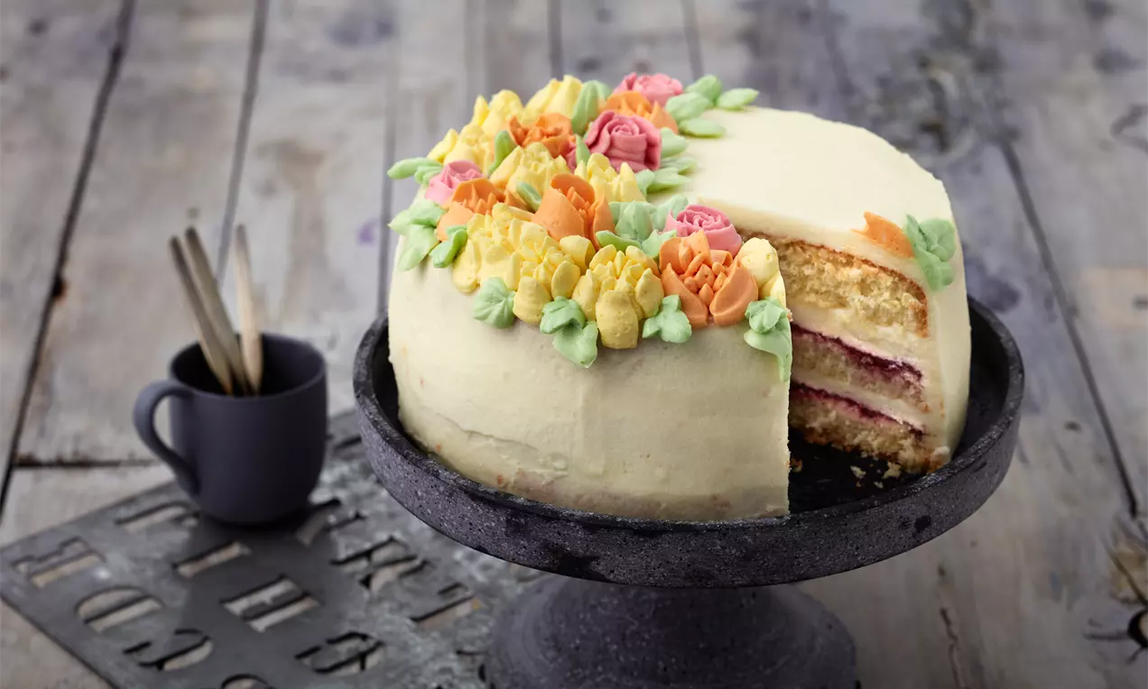 Helle Mascarpone-Torte mit bunter Blüten-Dekoration, angeschnitten auf einer dunklen Tortenplatte, darunter ein Holzuntergrund