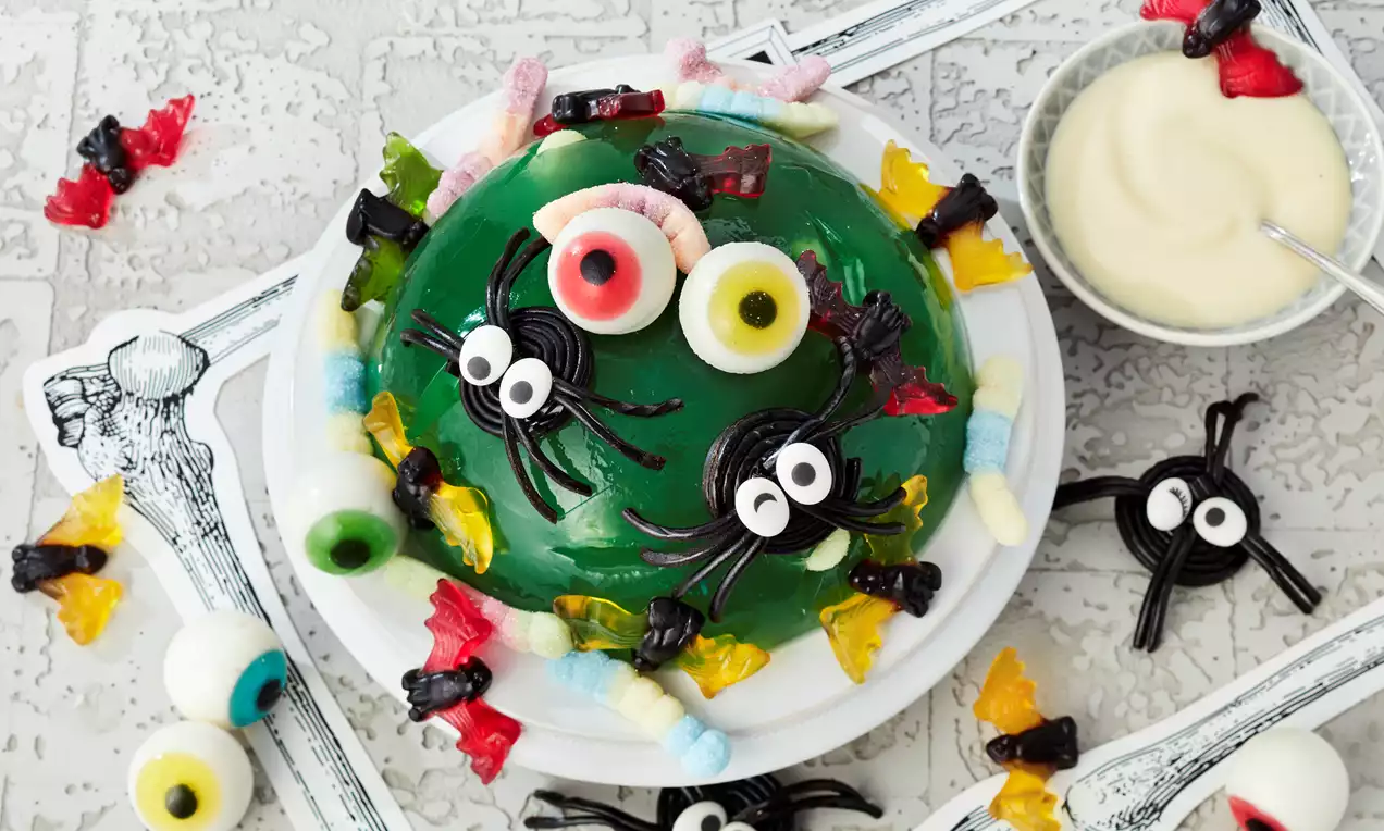 Grüner Wackelpudding in Halbkugel-Form, dekoriert mit Lakritz-Spinnen, bunten Gummisüßigkeiten und Augendekorationen auf weißem Teller