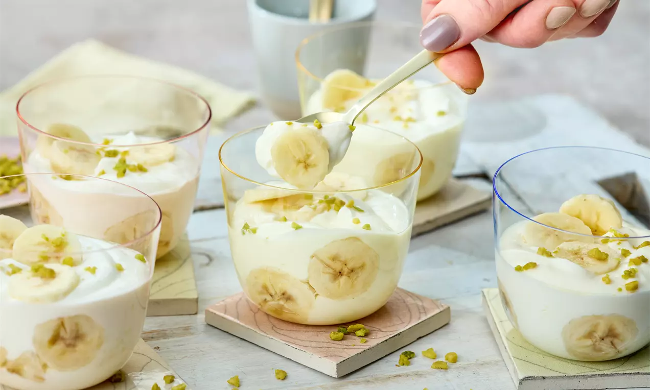 Gläser mit cremigem Quark, Bananenscheiben und gehackten Pistazien, angerichtet auf Holzuntersetzern, eine Hand mit Löffel greift zu