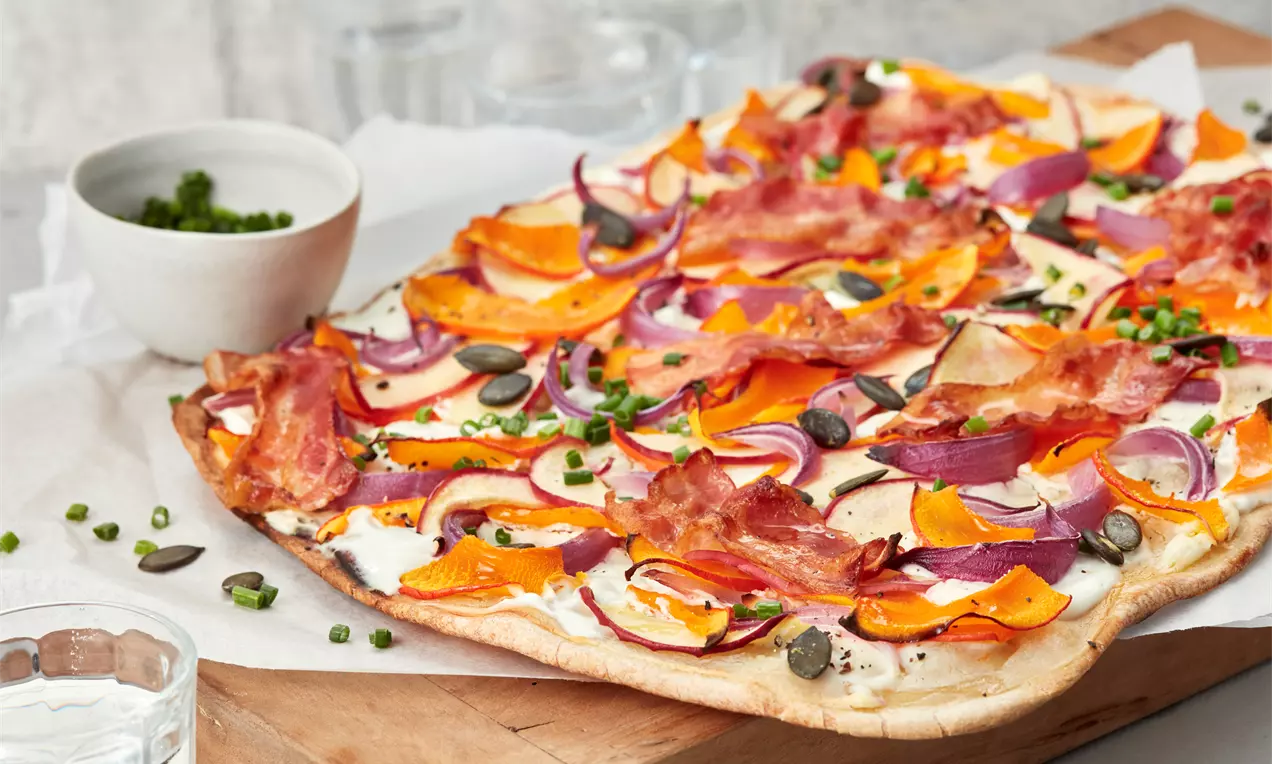 Rechteckiger Flammkuchen mit dünnem Boden, belegt mit orangefarbenem Kürbis, rotem Zwiebel, knusprigem Bacon und grünen Kürbiskernen