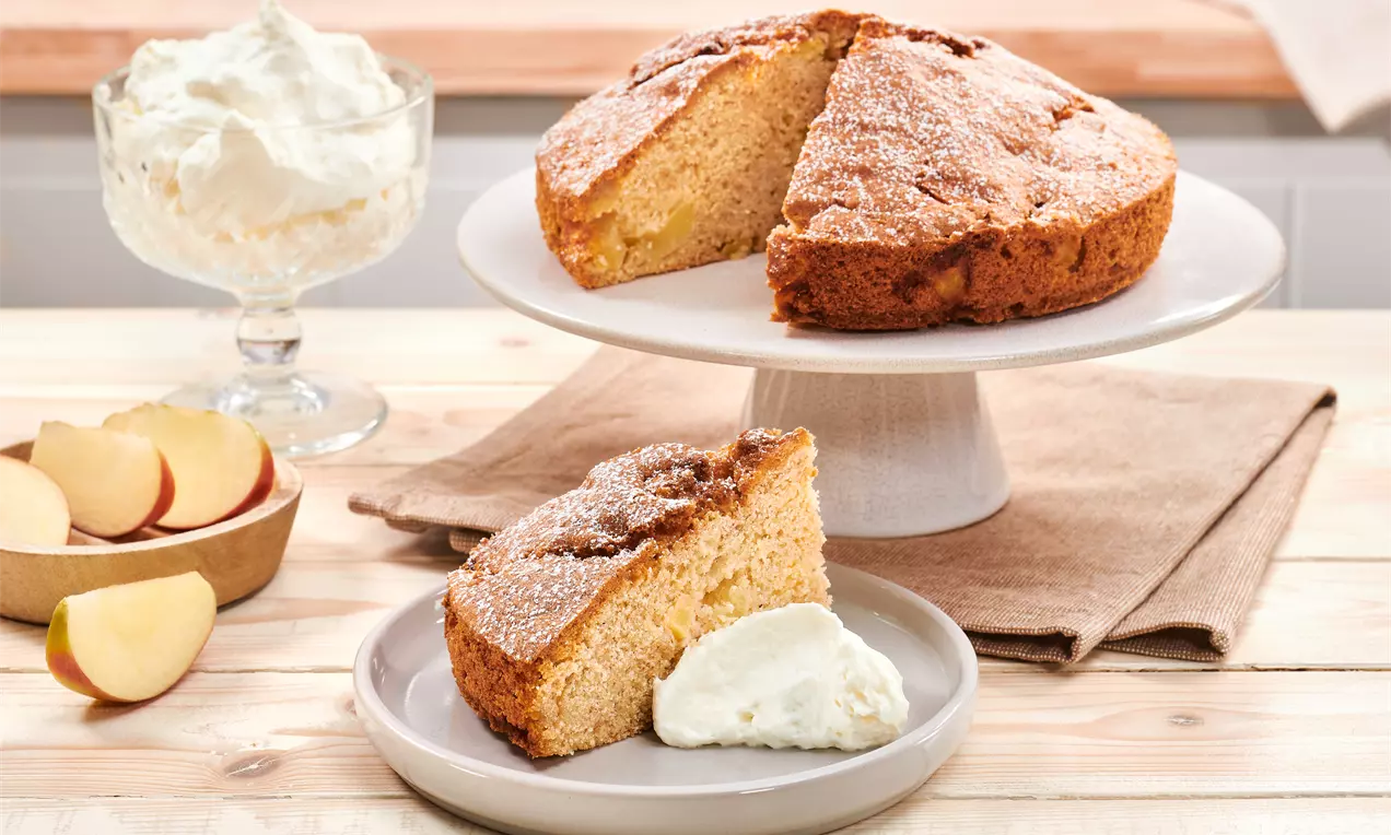 Apfelkuchen mit goldbrauner, leicht rissiger Kruste auf Tortenplatte, ein Stück mit Sahne auf Teller, dahinter Apfelspalten und Sahneschale