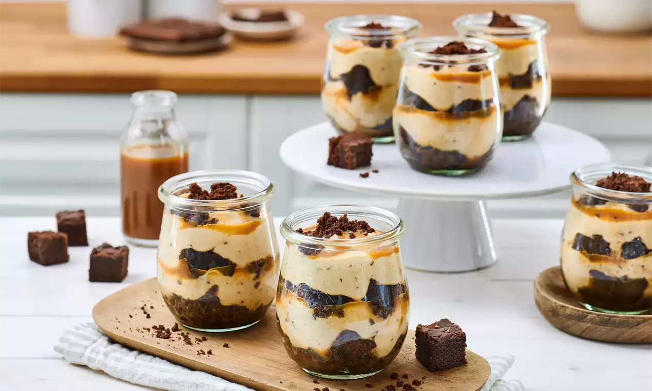 Geschichtete Schoko-Karamell-Creme in Gläsern, mit dunklen Brownie-Stückchen garniert, auf Holzbrett und Tortenplatte arrangiert