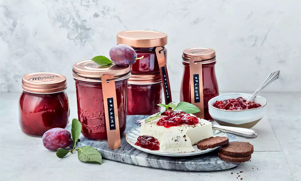 Pflaumen-Chutney in gläsernen, kupferfarben verschlossenen Einmachgläsern neben Käse mit rotem Chutney und frischen Pflaumen auf Steinplatte