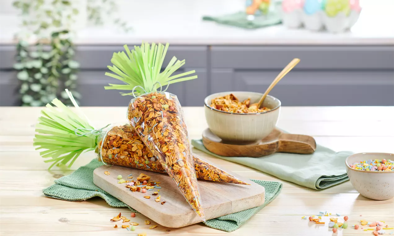 Zwei spitz zulaufende transparente Tüten in Form von Karotten, gefüllt mit orangefarbenem Granola, dekoriert mit grünem Papier auf Holzbrett