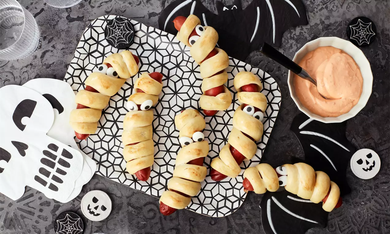 Würstchen in hellgoldenem Teig eingewickelt, mit Augen dekoriert, auf gemustertem Tablett und Halloween-Motiven wie Fledermäusen und Totenköpfen drapiert