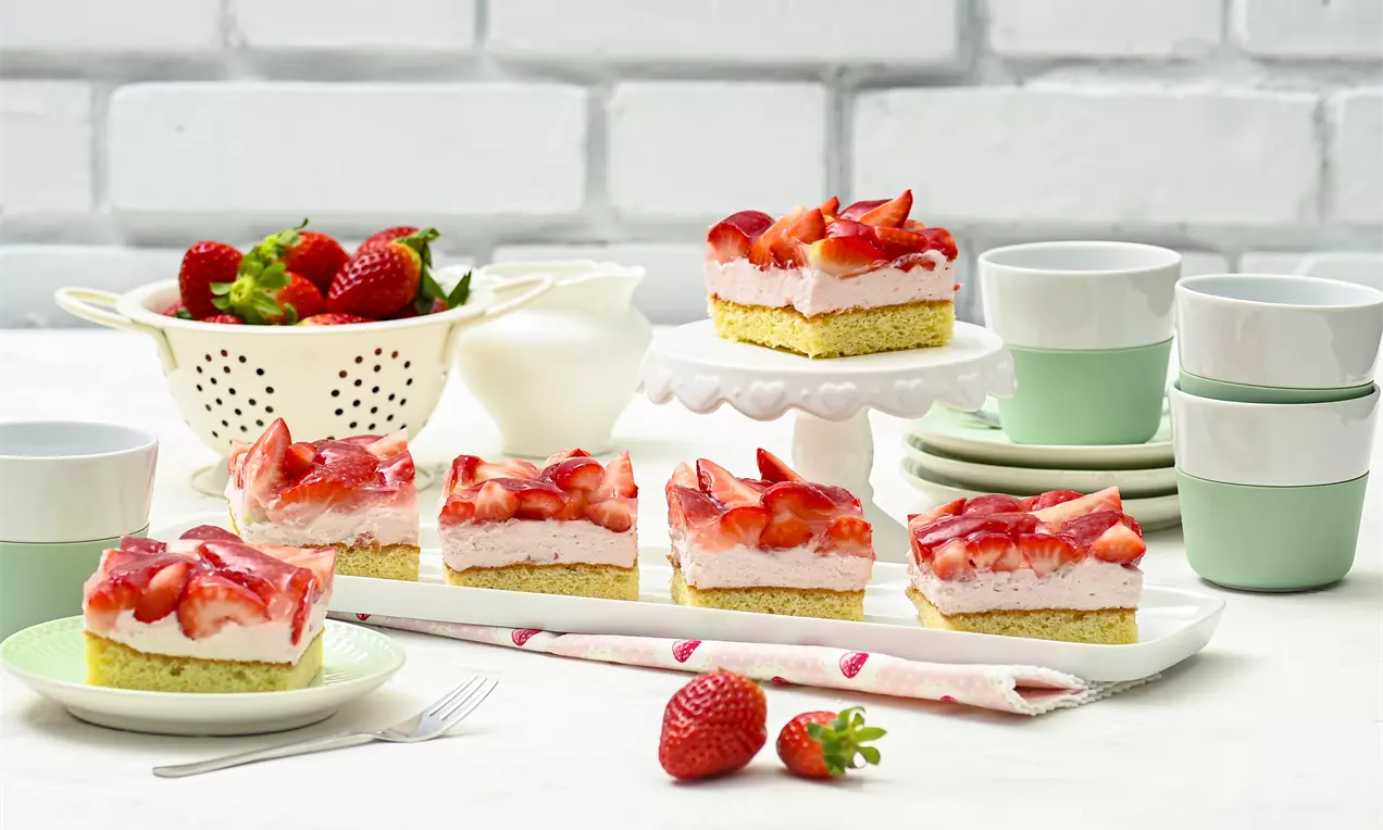 Erdbeerschnitten mit hellgelber Kuchenbasis, rosafarbener Creme und glänzender roter Erdbeerschicht auf weißer Platte mit frischen Erdbeeren