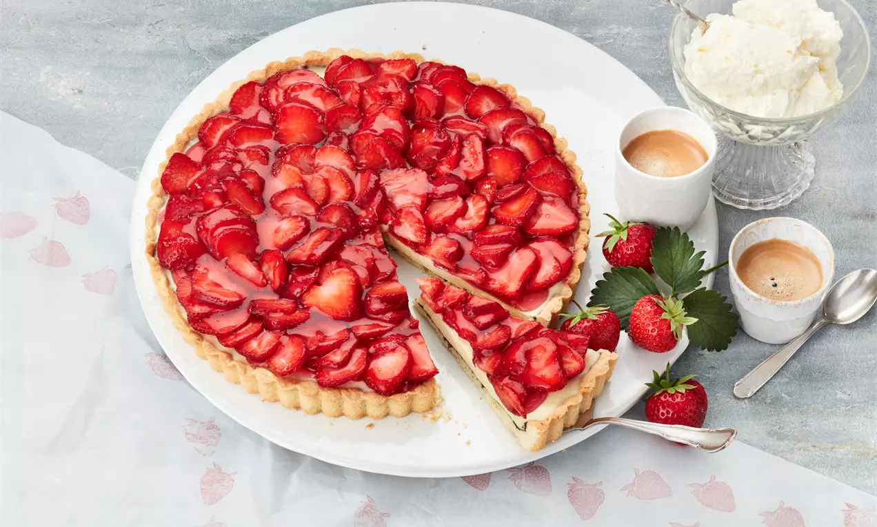 Erdbeertarte mit goldbraunem Mürbeteigboden, dicht belegt mit glänzenden roten Erdbeerscheiben auf weißem Teller mit frischen Erdbeeren dekoriert