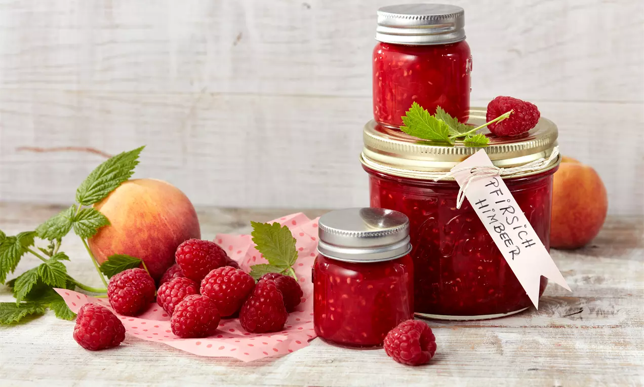 Gläser mit tiefroter Pfirsich-Himbeer-Konfitüre, dekoriert mit frischen Himbeeren, Pfirsichen und grünem Blatt auf hellem Holztisch