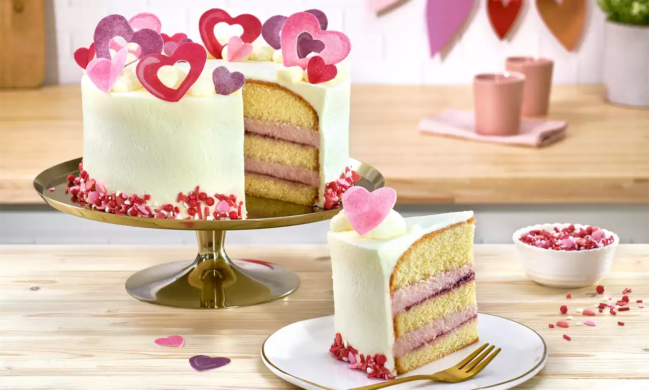 Valentinstag-Torte mit weißer Creme, rosa Fruchtschichten, Herzdekorationen aus Zucker und Streudekor, auf goldenem Tortenständer