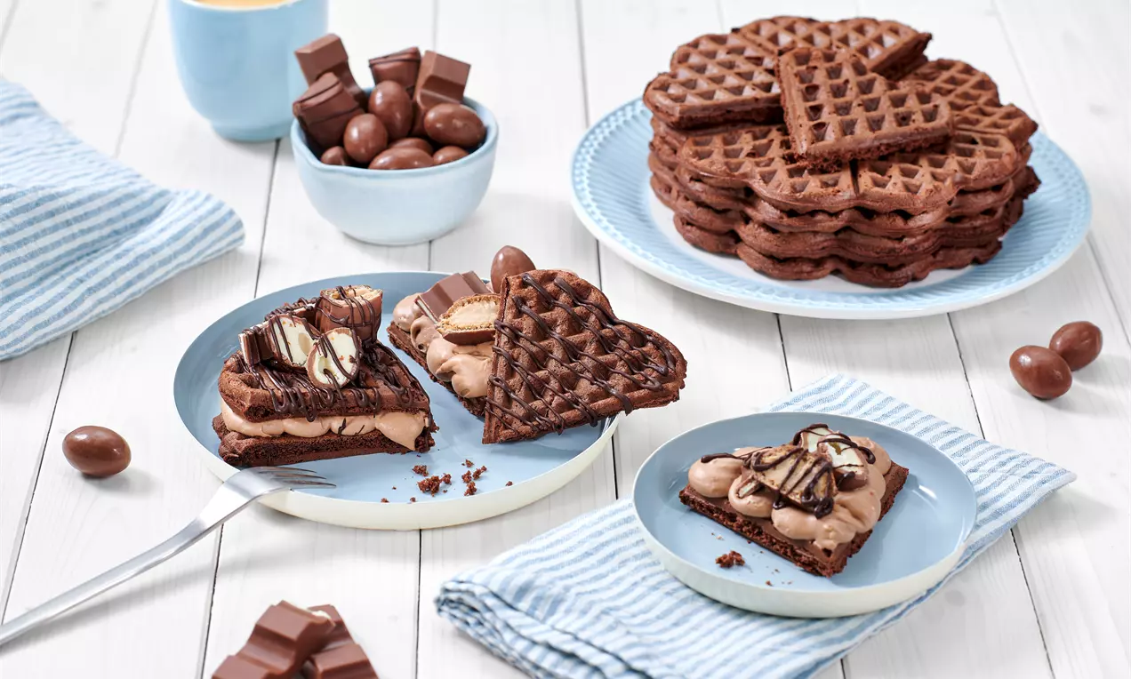 Schokowaffeln in Herzform, dunkelbraun und weich, teils mit cremiger Schokofüllung und Dekoration, arrangiert auf Tellern neben Kaffeebecher