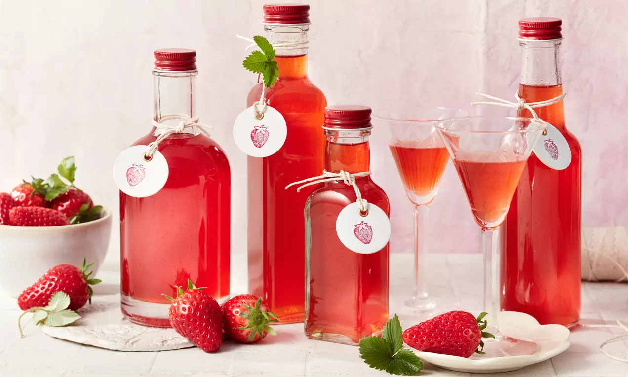 Erdbeerlikör in klarem Glas mit tiefroter Flüssigkeit, frischen halbierten Erdbeeren, Minzblatt und Juteband am Flaschenhals
