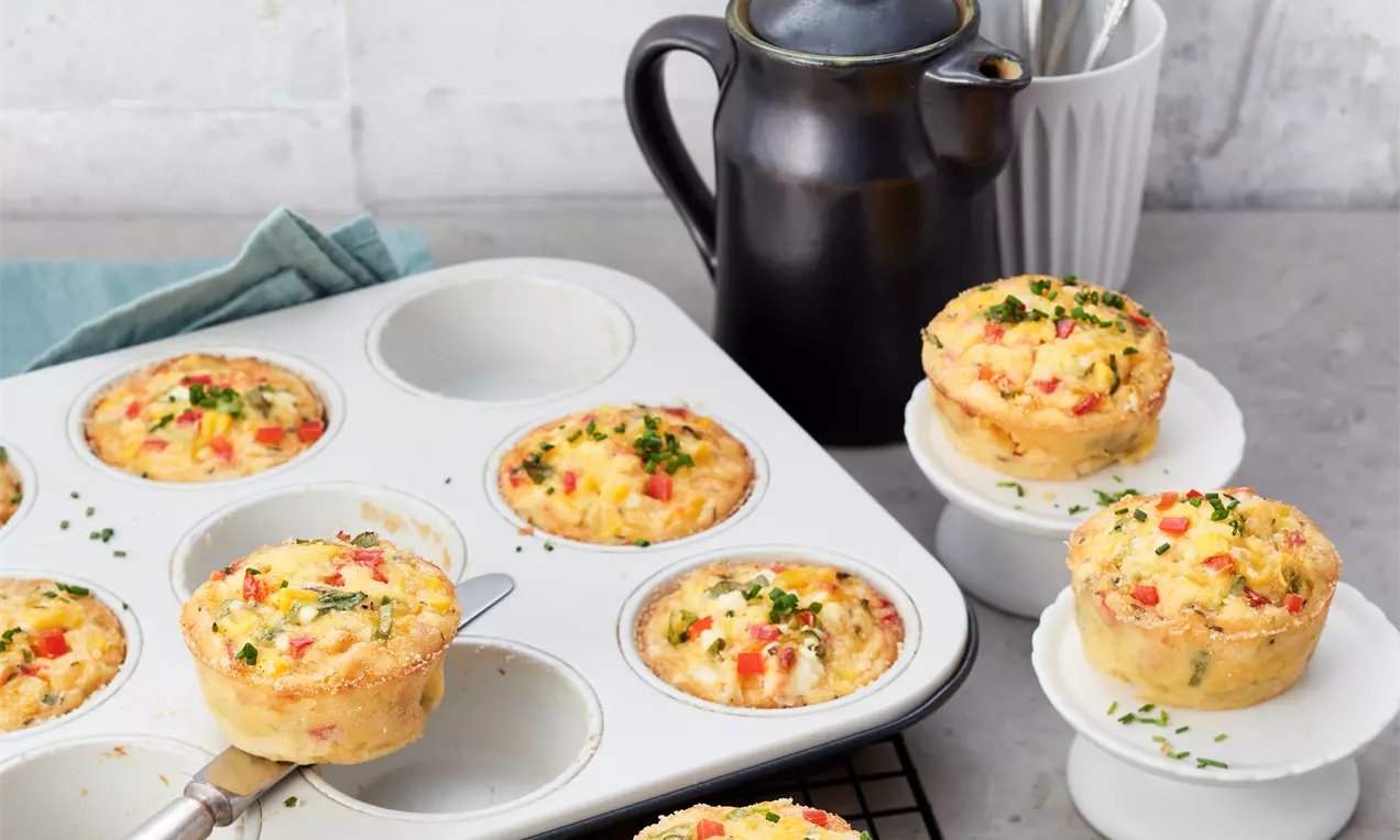 Goldbraune, luftige Rührei-Muffins mit roten Paprikastückchen und grünem Schnittlauch, teils in einer Backform, teils auf weißen Tellern angerichtet