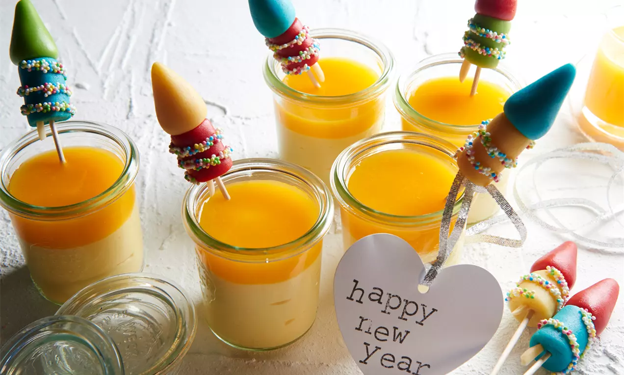 Marzipan-Pudding mit blassgelber Textur, glänzender Mandarinensoße und bunten Raketen-Dekospießen in Gläsern auf weißem Untergrund