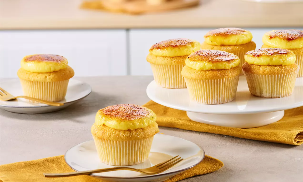 Goldgelbe Creme Brulée Cupcakes mit karamellisierter Oberfläche in Papierförmchen, arrangiert auf weißer Tortenplatte und Tisch mit gelbem Tuch
