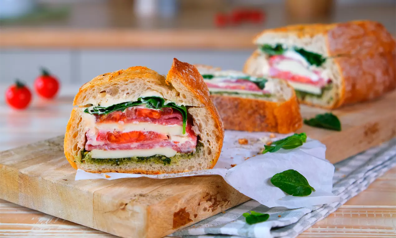 Rustikales Brot gefüllt mit Schichten aus roten Tomaten, grünem Spinat, Käse und Salami auf Holzbrett mit Butterbrotpapier
