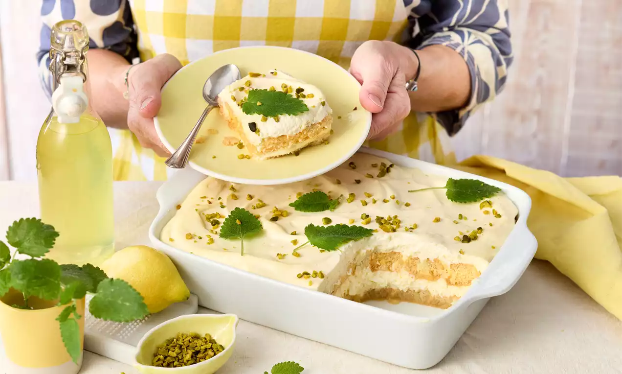 Limoncello-Tiramisu mit hellgelber Creme und grünem Melissenblatt in weißer Form, daneben Zitrone, Pistazien und Limoncello-Flasche