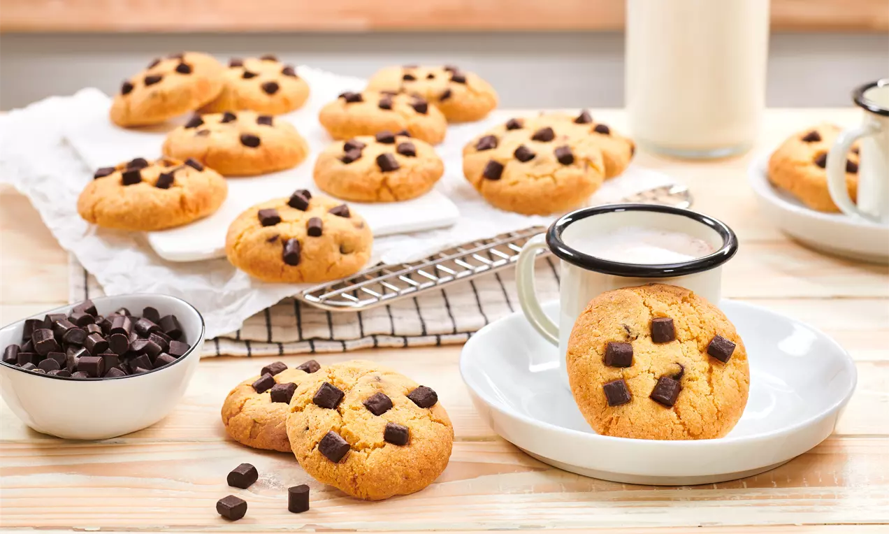 Goldbraune Cookies mit dunklen Schokostückchen auf Holzplatte, teilweise auf weißem Teller mit Emaille-Tasse und Schoko in kleiner Schale